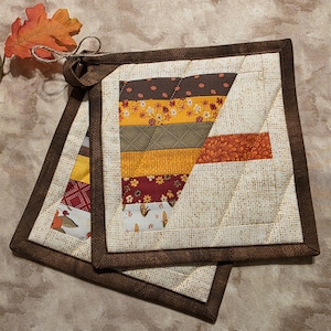 May include: Two square quilted pot holders featuring a patchwork design of autumn-colored fabric strips in shades of brown, orange, yellow, and cream. The pot holders have a dark brown fabric border and a small fabric loop for hanging.