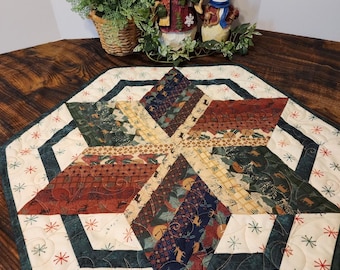May include: A green, white, and red quilted table runner with a snowflake design. The runner is made of fabric scraps and has a starburst pattern in the center. The runner is on a wooden table with two snowman figurines in the background.