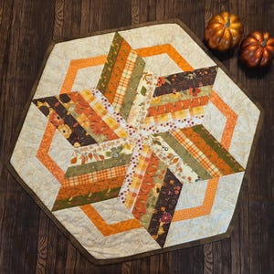 May include: A handmade, star-shaped quilted table topper with a beige background and a brown border. The star design features strips of fabric in shades of orange, brown, green, and yellow. Two small orange pumpkins sit on the wooden surface.