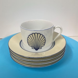 May include: A white teacup with a blue seashell design and a matching saucer. The teacup is placed on a stack of three additional saucers. The saucers are cream-colored with a blue trim.