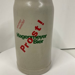 May include: A white ceramic beer stein with a red and green logo that says "Prost! Hagenmeyer Bier" and the text "immer wieder" in red.