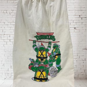 May include: Off-white drawstring bag featuring the Teenage Mutant Ninja Turtles. The graphic shows the four turtles in their signature colors, holding weapons. The bag is hanging on a white hanger against a white brick wall.