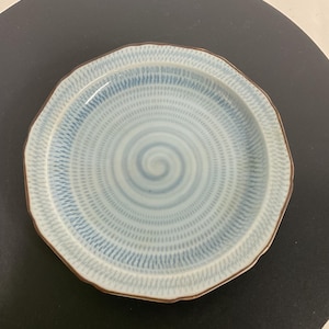 May include: A ceramic plate with a light blue spiral design and a brown rim. The plate has an irregular, wavy edge. The plate is on a black surface.