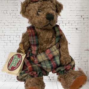 May include: A brown teddy bear wearing a plaid cap and overalls. The bear has a black nose and is holding a tag that reads "Cottage Collectibles Artist Designed". The overalls are red, green, and tan plaid.