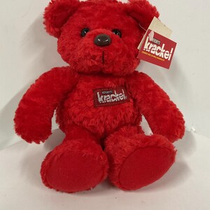 May include: Red plush teddy bear with a Hershey's Krackel chocolate bar logo on its chest. The bear has black eyes and a brown nose.