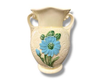 May include: Cream-colored ceramic vase with two handles and a flared rim. The vase features a raised blue flower with green leaves and a textured, swirling pattern. A decorative piece for home decor.