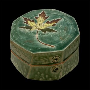 May include: A green ceramic hexagonal box with a detailed maple leaf design on the lid. The leaf features shades of brown and yellow. The box has two small circular clasps and a textured surface.