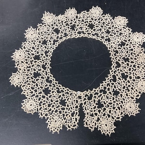 May include: Cream-colored lace collar with a circular design. The collar features intricate floral patterns and a delicate, openwork texture. The collar is designed to be worn around the neck and is likely a vintage or handmade item.