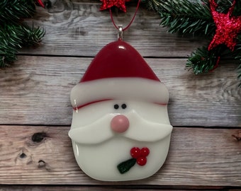 Könnte beinhalten: Ein Weihnachtsschmuck aus geschmolzenem Glas in Form eines Weihnachtsmanns mit roter Mütze und weißem Rand, weißem Schnurrbart, rosa Nase, zwei schwarzen Augen und einem grünen Stechpalmenblatt mit drei roten Beeren.