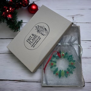 May include: A green and red glass Christmas wreath ornament in a white box with a white and grey tissue paper bag. The box has the text "FRIT & FLAME HANDMADE GLASS ART & JEWELLERY" on it.