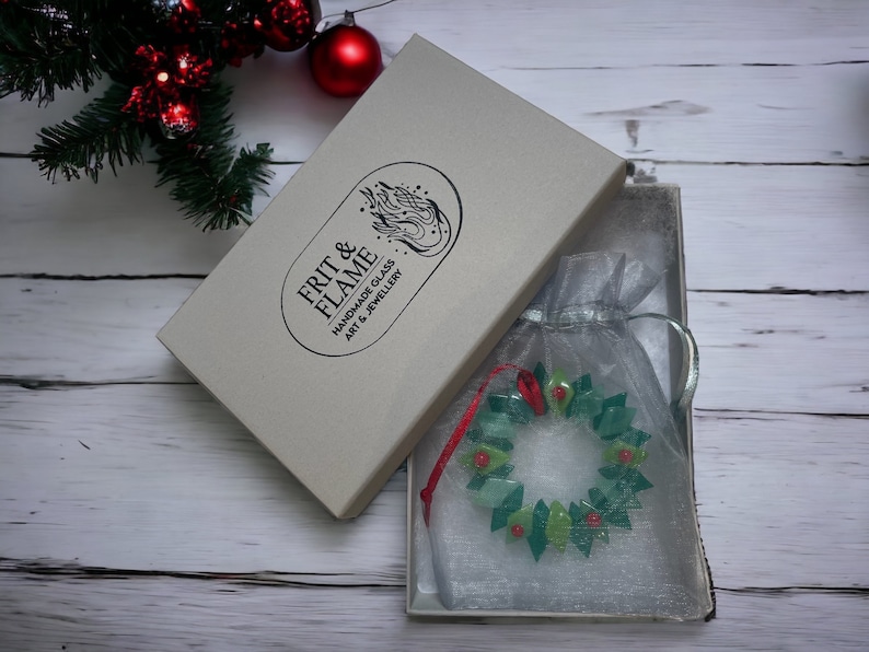 May include: A green and red glass Christmas wreath ornament in a white box with a white and grey tissue paper bag. The box has the text "FRIT & FLAME HANDMADE GLASS ART & JEWELLERY" on it.