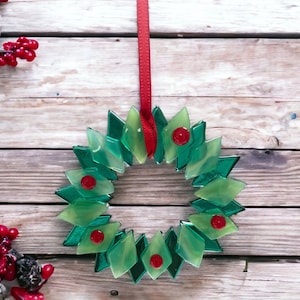 May include: A green glass Christmas wreath with red accents hanging from a red ribbon on a wooden background.
