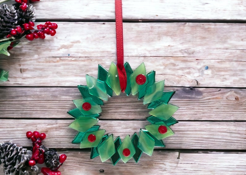 May include: A green glass Christmas wreath with red accents hanging from a red ribbon on a wooden background.