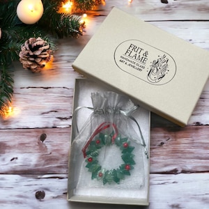 May include: A green glass Christmas wreath ornament with red berries, packaged in a white box with the text "FRIT & FLAME HANDMADE GLASS ART & JEWELLERY"
