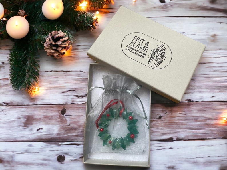 May include: A green glass Christmas wreath ornament with red berries, packaged in a white box with the text "FRIT & FLAME HANDMADE GLASS ART & JEWELLERY"