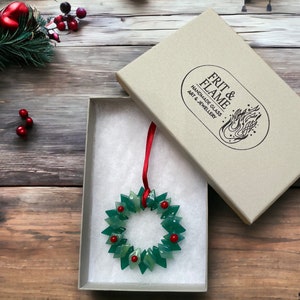 May include: A green glass Christmas wreath ornament with red glass berries, in a white gift box with a red ribbon. The box is labelled "FRIT & FLAME HANDMADE GLASS ART & JEWELLERY"