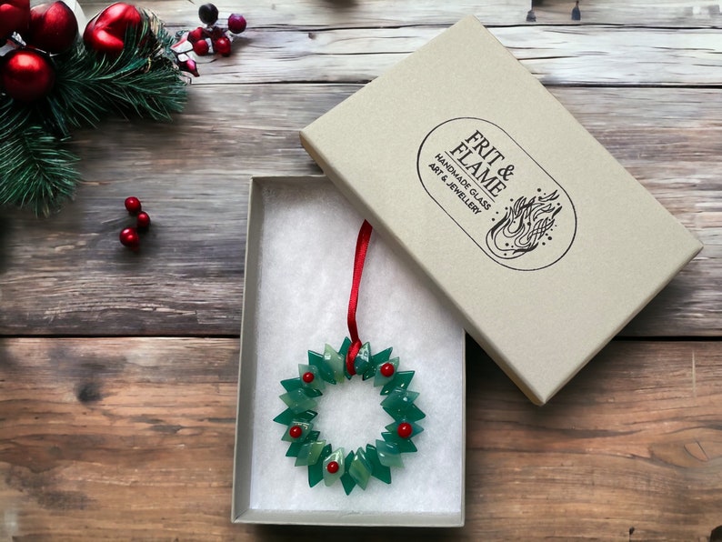 May include: A green glass Christmas wreath ornament with red glass berries, in a white gift box with a red ribbon. The box is labelled "FRIT & FLAME HANDMADE GLASS ART & JEWELLERY"