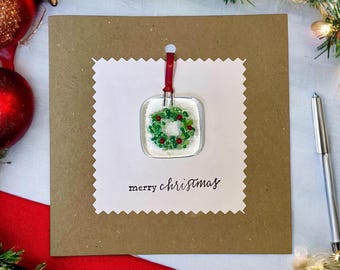 May include: A square greeting card made of brown cardstock with a white, scalloped-edge insert. The insert features a fused glass ornament shaped like a wreath with green and red beads, suspended by a red ribbon. The text merry christmas is printed.