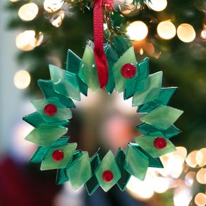 May include: A green glass Christmas bauble shaped like a wreath with red berries. The bauble is hanging from a red ribbon on a Christmas tree.