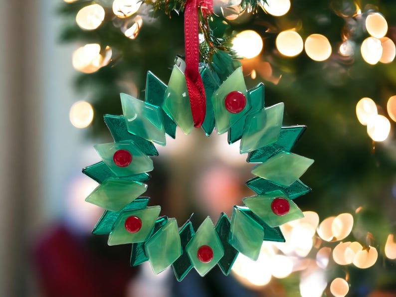 May include: A green glass Christmas bauble shaped like a wreath with red berries. The bauble is hanging from a red ribbon on a Christmas tree.