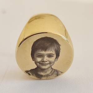 Könnte beinhalten: Ein goldfarbener Siegelring mit einem eingravierten Porträt eines Kindes auf der flachen Oberfläche. Der Ring hat eine glatte, polierte Oberfläche und eine abgerundete Form. Das Porträt ist in Schwarzweiß gehalten.