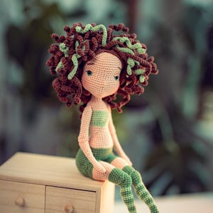 Op de afbeelding: Een handgemaakte gehaakte pop met bruin en groen haar, gekleed in een groene en roze outfit. De pop zit op een kleine houten ladekast. Het haar van de pop is een mix van bruin en groen garen, en de outfit bestaat uit een gestreepte top en shorts.