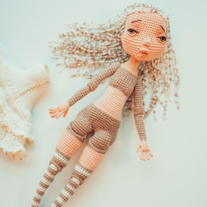 Puede incluir: Muñeca de ganchillo con cabello largo y castaño claro y una expresión neutra. La muñeca lleva un atuendo bicolor marrón y melocotón con piernas a rayas. Un vestido de ganchillo con detalles de cuentas es visible en el fondo.