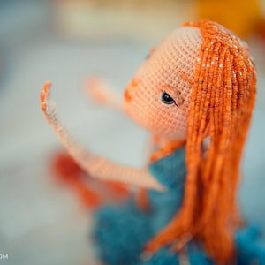 Puede incluir: Muñeca hecha a mano con cabello naranja y vestido azul. El cabello de la muñeca está hecho de pequeñas cuentas naranjas. La muñeca está tejida a ganchillo y tiene un tono de piel claro. La muñeca está sentada con un brazo levantado.
