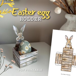 Puede incluir: Porta huevos de Pascua imprimible con un conejo y una caja de madera. El soporte está diseñado para ser ensamblado a partir de una plantilla PDF. El texto "Easter egg holder" y "PDF Printable" es visible en la imagen.