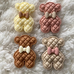May include: Four teddy bear-shaped embellishments in pastel shades: yellow, pink, brown, and beige. Each bear features a quilted design and a contrasting bow tie. These decorative elements are suitable for crafts and DIY projects.