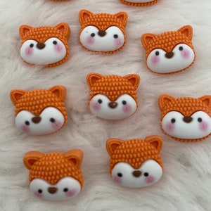Puede incluir: Diez botones de color naranja en forma de zorro con caras blancas y mejillas rosadas. Los botones tienen una superficie texturizada.