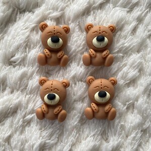 Puede incluir: Cuatro pequeños adornos de osos de peluche marrones con narices negras, vientres color crema y detalles cosidos. Los osos están dispuestos sobre una superficie blanca texturizada, creando una estética encantadora y juguetona.