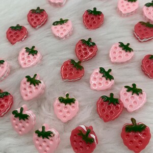 Può includere: Una collezione di bottoni a forma di fragola rosa e rossa con foglie verdi. I bottoni sono disposti su una superficie bianca e soffice.