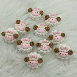 May include: A collection of ten sheep-shaped embellishments. Each sheep has a white, textured body, a pink face, and brown curly horns. A pink bow adorns the top of each sheep's head. The embellishments are arranged on a white, fluffy surface.