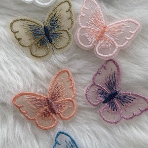 Könnte beinhalten: Sechs gestickte Schmetterlingsapplikationen in verschiedenen Farben, darunter Weiß, Silber, Rosa, Beige und Blau. Die Schmetterlinge haben einen zarten, durchsichtigen Stoff und aufwendige Stickdetails.