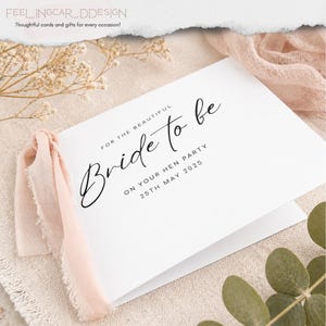 May include: A white folded greeting card featuring black text that reads FOR THE BEAUTIFUL Bride to be ON YOUR HEN PARTY 25TH MAY 2025. The card is adorned with a soft, light pink frayed fabric ribbon attached to the left side of the card cover.