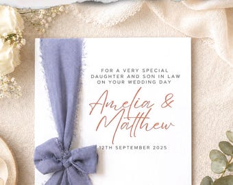 Könnte beinhalten: Eine quadratische weiße Hochzeitskarte mit einem staubblauen, ausgefransten Band, das auf der linken Seite zu einer Schleife gebunden ist. Der Text lautet: FOR A VERY SPECIAL DAUGHTER AND SON IN LAW ON YOUR WEDDING DAY, Amelia & Matthew, 12TH SEPTEMBER 2025 in Schwarz und Kupfer.