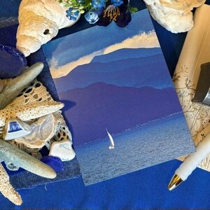 May include: A rectangular postcard featuring a blue-toned landscape of layered mountains and a small sailboat on the water. The card is surrounded by white seashells, a starfish, a white pen, and a postage stamp showing an American flag.