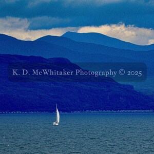 May include: A white sailboat floats on deep blue water in front of dark blue mountain ranges under a cloudy sky. The text K. D. McWhitaker Photography © 2025 is centered over the landscape. The scene captures a calm maritime view.