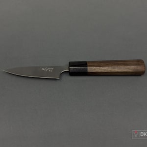 May include: A small, stainless steel knife with a dark wooden handle. The blade has a pointed tip and a small etched design. The handle is a combination of dark and light wood tones. The knife is set against a gray background.