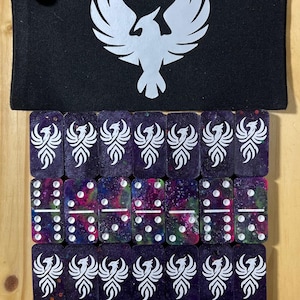 May include: A black zippered pouch with a silver phoenix logo and a skull charm. Below, dominoes with a phoenix design and colorful galaxy patterns are arranged on a wooden surface.