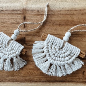 May include: Two handmade macrame ornaments with cinnamon sticks and wooden beads. The ornaments are white and have a fringe around the bottom.