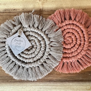 May include: Two handmade macrame coasters, one in a light beige colour and the other in a dusty rose colour. Both coasters have a circular design with a fringe border. A small tag with the text "Creative Craft Studio" is attached to the beige coaster.