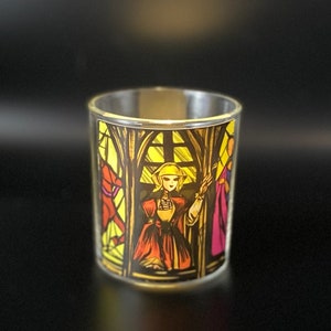 Peut inclure: Un photophore en verre transparent avec un motif de vitrail. Le motif représente une femme en robe rouge et orange, avec des accents jaunes et dorés. Le fond est une fenêtre en vitrail jaune.