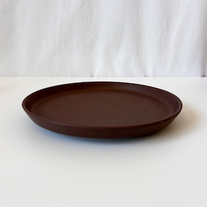May include: A circular, dark brown plate with a smooth, matte finish and a slightly raised rim. The plate features a simple, flat design with a consistent deep brown color throughout, presenting a clean and functional aesthetic for serving food.
