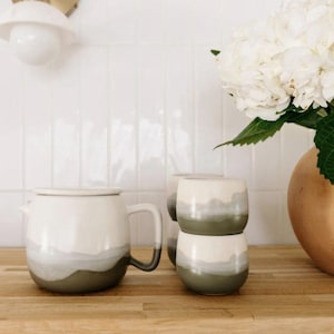 May include: A white and gray ceramic teapot with a matching lid and handle, paired with two stacked ceramic cups. The cups and teapot feature a layered design in white, gray, and olive green. The set is displayed on a wooden surface.