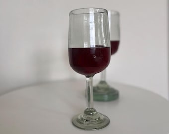 May include: Two clear wine glasses, each containing a red liquid, possibly wine, stand on a white surface. The glasses have a rounded bowl and a thin stem. The background is a plain white wall.