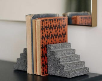May include: A pair of grey, speckled stone bookends shaped like miniature staircases, holding three vintage books. The front book features a vibrant orange cover with a repeating black geometric pattern of stylised bird-like figures.