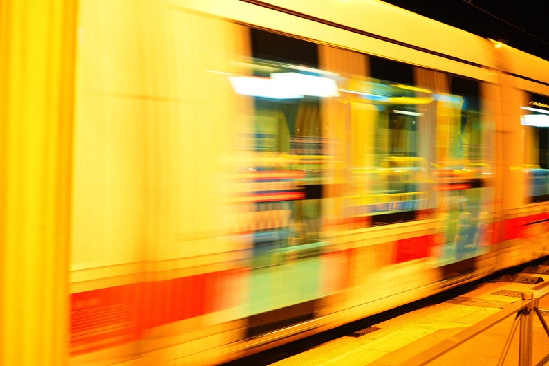 Op de afbeelding: Een wazig beeld van een gele trein die snel door een stad rijdt in de nacht. De ramen van de trein zijn verlicht en de lichten reflecteren op de buitenkant van de trein.