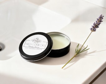 May include: A round black metal tin containing a pale yellow cleansing balm, with the lid placed beside it. The lid features a white label with the text Wild Comfort Cleansing Balm and a floral illustration. A sprig of purple lavender lies nearby.
